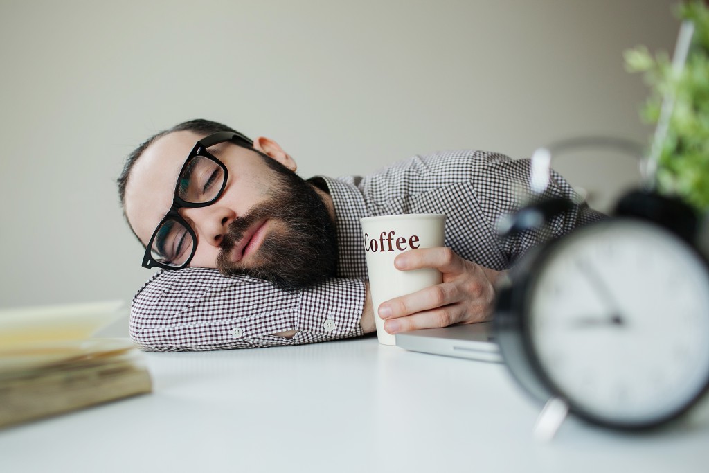 Man with beard in glasses sleeps in office on the table over laptop with coffee in hand