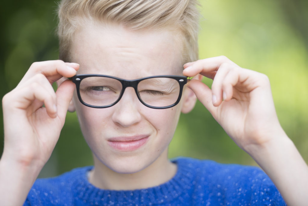 Portrait smart looking blond teenager with glasses and a blink of an eye, thoughtful and clever, outdoor with green blurred background.