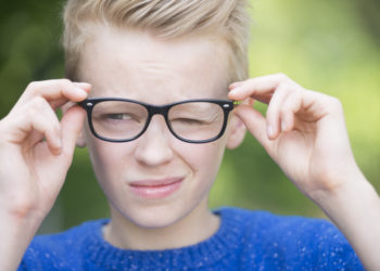 Portrait smart looking blond teenager with glasses and a blink of an eye, thoughtful and clever, outdoor with green blurred background.