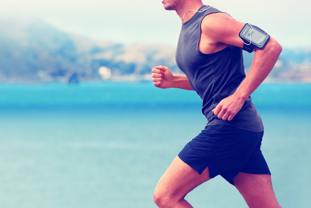 Cardio runner running listening smartphone music. Unrecognizable body jogging on ocean beach or waterfront working out with heart rate monitor app device and earphones in summer.