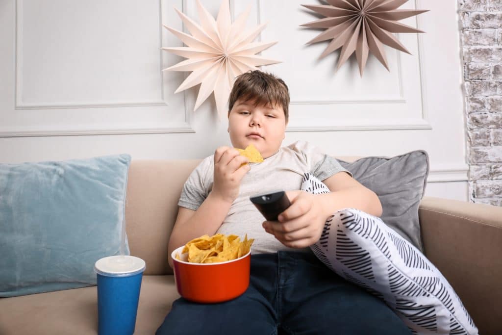 Ein Junge sitzt vorm Fernseher, isst Chips und trinkt Cola.