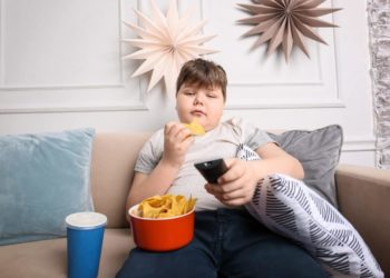 Ein Junge sitzt vorm Fernseher, isst Chips und trinkt Cola.