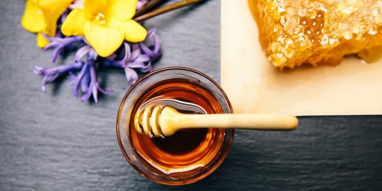 Honig in Glasschale mit Holzlöffel neben Honigwabe und losen Schnittblumen.