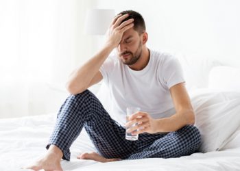 Mann sitzt im Bett und fasst sich mit einer Hand an den schmerzenden Kopf und hält in der anderen Hand ein Glas mit Wasser