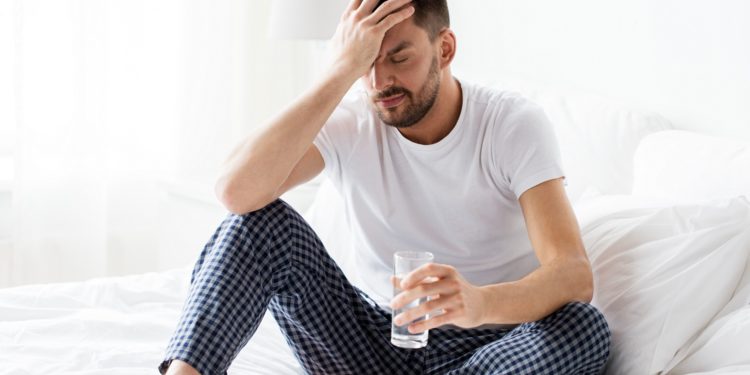 Mann sitzt im Bett und fasst sich mit einer Hand an den schmerzenden Kopf und hält in der anderen Hand ein Glas mit Wasser