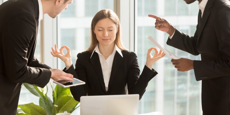 Eine Frau sitzt im Büro und meditiert, während zwei Männer auf sie einreden.