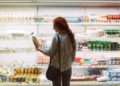 Junge Frau steht vor Kühlregal im Supermarkt und betrachtet Milchflasche in ihrer Hand