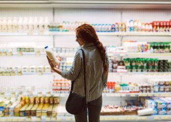 Junge Frau steht vor Kühlregal im Supermarkt und betrachtet Milchflasche in ihrer Hand