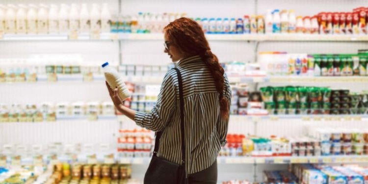Junge Frau steht vor Kühlregal im Supermarkt und betrachtet Milchflasche in ihrer Hand