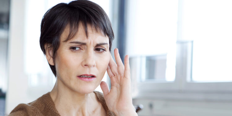 Eine Frau berührt ihr Ohr mit der Hand und zeigt einen gestressten Gesichtsausdruck.