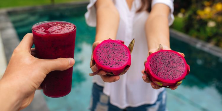 Ein Smoothie mit Drachenfrucht sowie zwei Hälften einer aufgeschnittenen Drachenfrucht in den Händen einer Frau