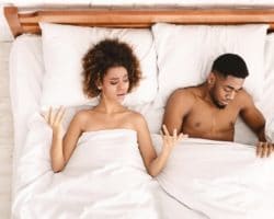 Junges Paar liegt im Bett, Frau hebt Hände fragend, Mann schaut an sich unter der Bettdecke herunter