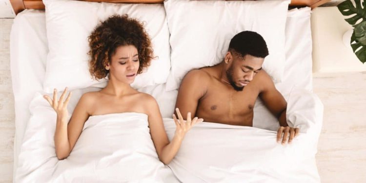 Junges Paar liegt im Bett, Frau hebt Hände fragend, Mann schaut an sich unter der Bettdecke herunter