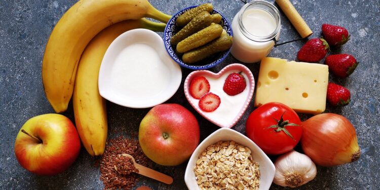 Verschiedene Lebensmittel wie Bananen, Joghurt, Äpfel, Käse, saure Gurken und Leinsamen vor grauem Hintergrund