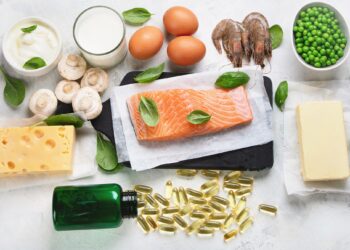 Vitamin-D-haltige Nahrungsmittel wie Lachs, Butter, Erbsen, Käse und Eier sowie ein Fläschchen mit ausgekippten Kapseln eines Nahrungsergänzungsmittels vor weißem Hintergrund
