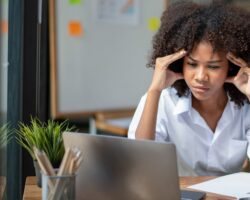 Frau sitzt am Schreibtisch und schaut angestrengt auf den Laptop, während sie sich mit den Händen die Schläfen hält