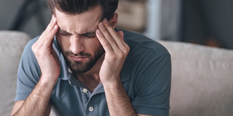 Mann in blauem Poloshirt massiert mit den Fingerspitzen beide Schläfen und verzieht das Gesicht vor Schmerzen