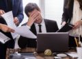 Mann sitzt gestresst im Büro umkreist von Mitarbeitenden.