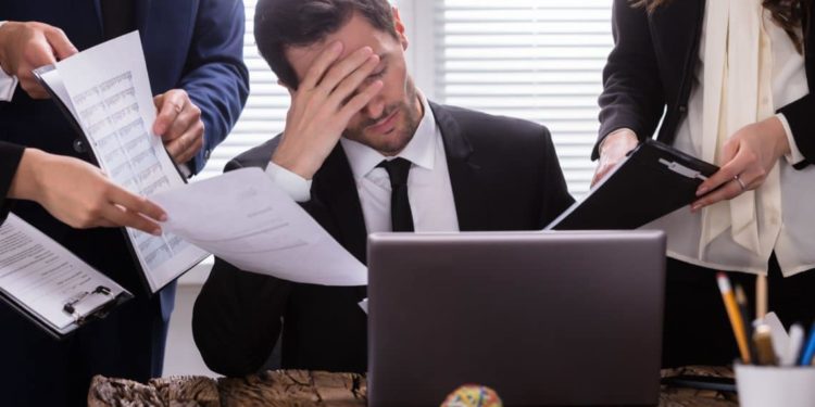 Mann sitzt gestresst im Büro umkreist von Mitarbeitenden.