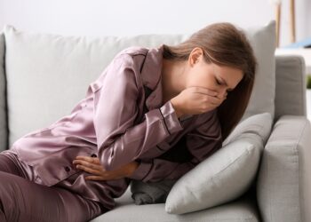 Frau liegt auf dem Sofa und hält sich vor Übelkeit die Hand vor den Mund.