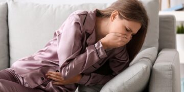 Frau liegt auf dem Sofa und hält sich vor Übelkeit die Hand vor den Mund.