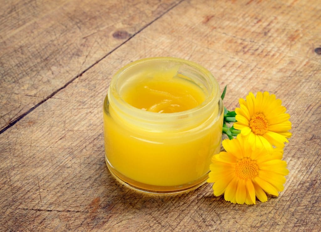 Gelbe Salbe in durchsichtigem Tiegel und zwei Ringelblumen auf einem Holztisch.