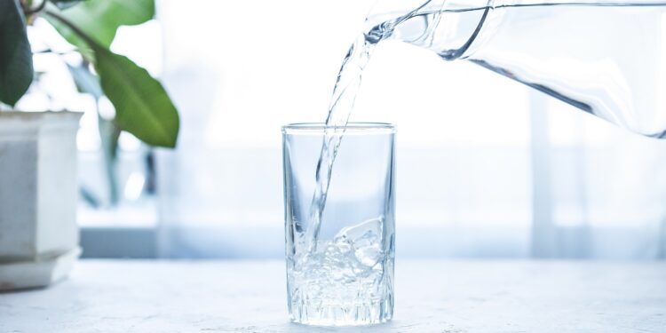 Aus einer gläsernen Wasserkaraffe fließt klares Wasser in ein Glas