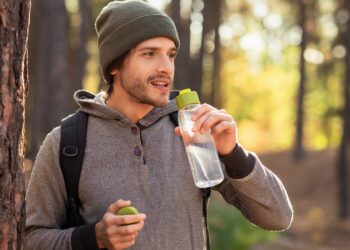 Mann mit Mütze und Rucksack im Wald hält eine Trinkflasche mit Wasser in der Hand