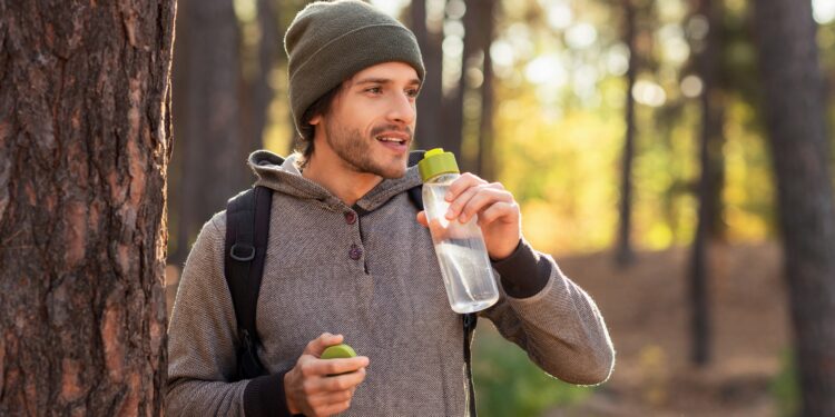 Mann mit Mütze und Rucksack im Wald hält eine Trinkflasche mit Wasser in der Hand