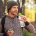 Mann mit Mütze und Rucksack im Wald hält eine Trinkflasche mit Wasser in der Hand