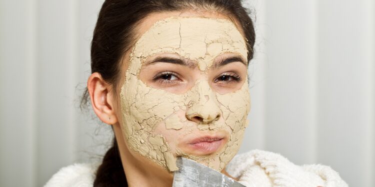 Eine Frau in weißem Bademantel kratzt sich mit einem Spachtel die Reste einer Kieselerde-Gesichtsmaske ab.