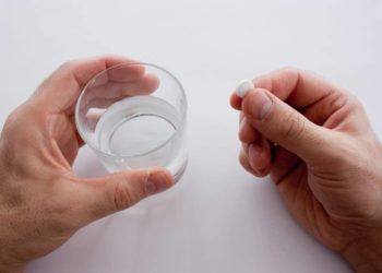 Eine Hand hält ein Wasserglas, die andere eine Tablette