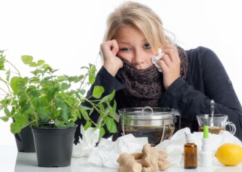 Erkältete Frau mit Taschentuch sitzt zwischen vielen verschiedenen naturheilkundlichen Mitteln