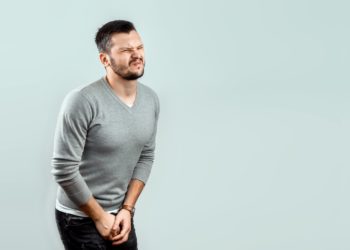 Ein Mann mit schmerzverzerrtem Gesicht hält sich die Hände vor den Genitalbereich.