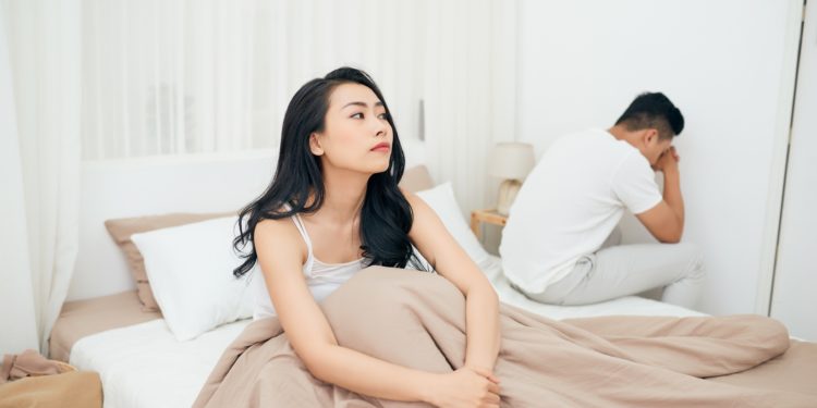 Ein Mann sitzt niedergeschlagen mit gesenktem Kopf auf der Bettkante, während seine Frau aufrecht im Bett sitzt und ratlos ins Leere schaut. 
