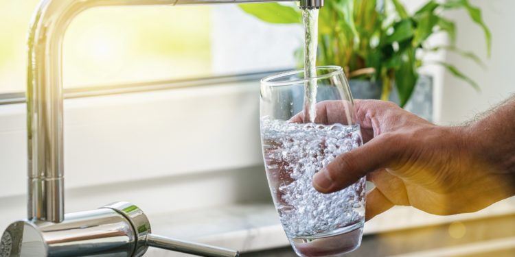 Ein Mann befüllt am Wasserhahn i der Küche ein Glas mit Leitungswasser. 