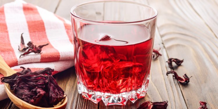 Auf einem Holztisch steht ein Glas Hibiskustee mit Eiswürfeln, daneben liegt ein rot-weiß kariertes, zusammengefaltetes Geschirrhandtuch und ein Holzlöffel mit getrockneten Hibiskusblüten. 