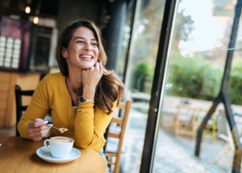 Eine Frau mit glücklichem Gesichtsausdruck sitzt an einem Fenster und trinkt Kaffee.