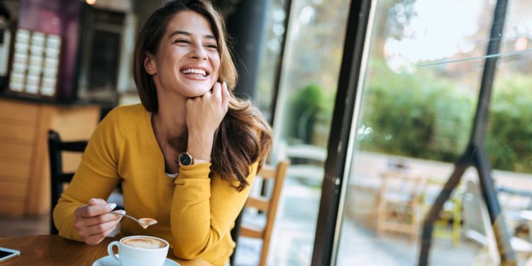 Eine Frau mit glücklichem Gesichtsausdruck sitzt an einem Fenster und trinkt Kaffee.