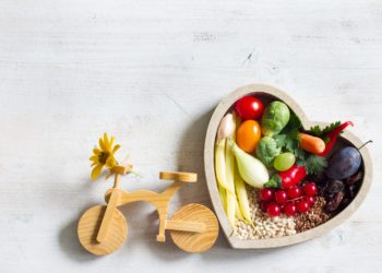 Herzfömige Schale gefüllt mit Obst und Gemüse. Links daneben ein Spielzeug Holzfahrrad.