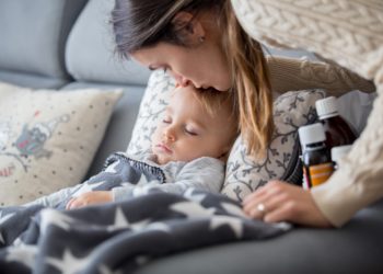 Eine Mutter küsst ihr Kind auf die Stirn, welches krank auf dem Sofa liegt und schläft.