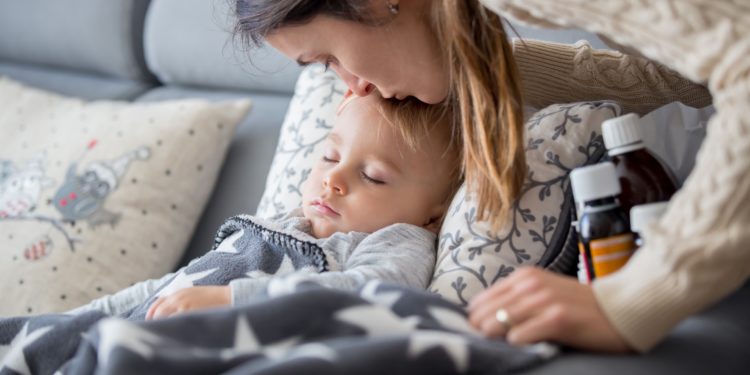 Eine Mutter küsst ihr Kind auf die Stirn, welches krank auf dem Sofa liegt und schläft. 