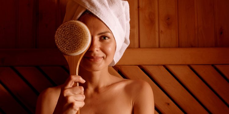 Frau mit weißem Handtuchturban sitzt in der Sauna und hält sich eine Massagebürste vor das rechte Auge.