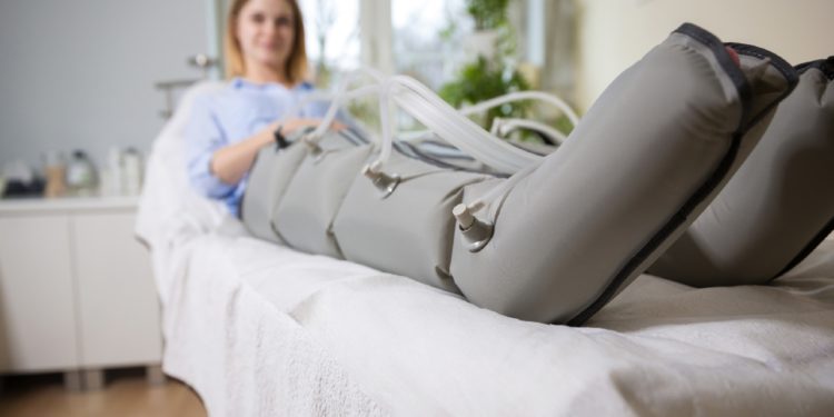 Eine Frau auf einer Liege hat an den Beinen die Druck-Manschetten eines Lymphdrainage-Geräts angelegt. 