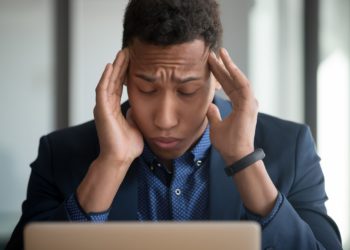 Ein erschöpft wirkender Man sitzt vor einem aufgeklappten Laptop und hält sich die Hände an die Schläfen.