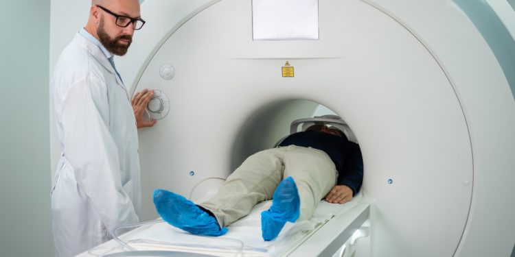 Ein Arzt nimmt Einstellungen an einem MRT-Gerät vor, in dem ein Mann bis zur Hüfte auf einer Liege liegt.
