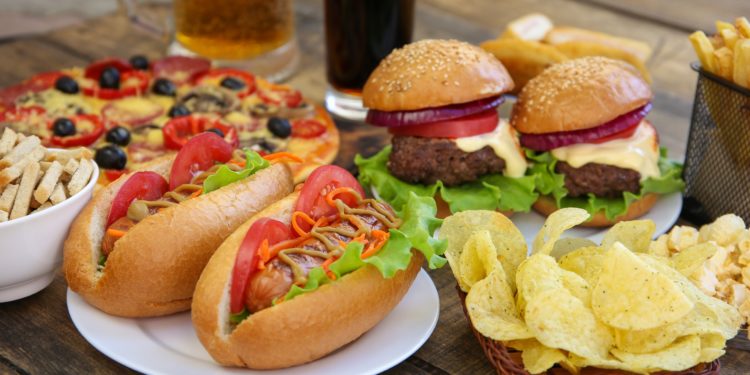 Burger, PIzza, Chips, Pommes, Hot Dogs, Cola und Bier auf einem Holztisch.