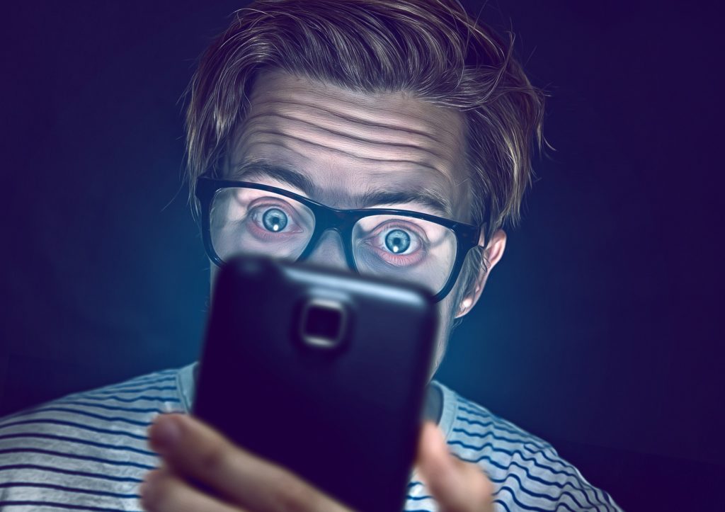A man wearing glasses stares at a smartphone with wide eyes.