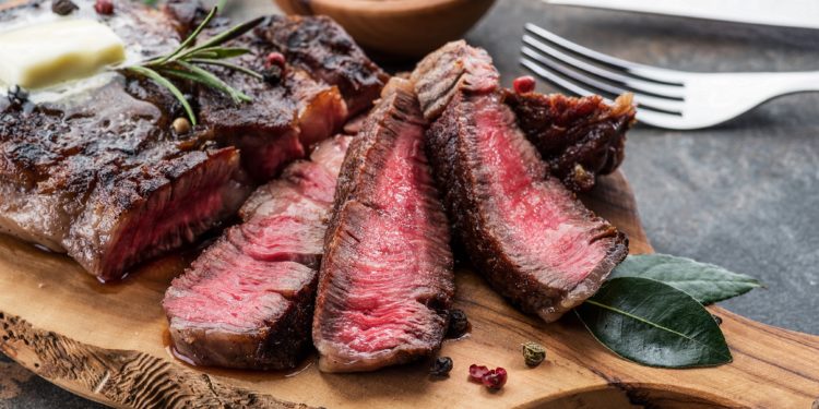 Medium rare Steak aufgenschitten auf einem Holzbrett mit Pfefferkörnern, Messer und Gabel daneben