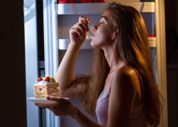 Eine Frau steht am Kühlschrank und isst ein Stück Kuchen.